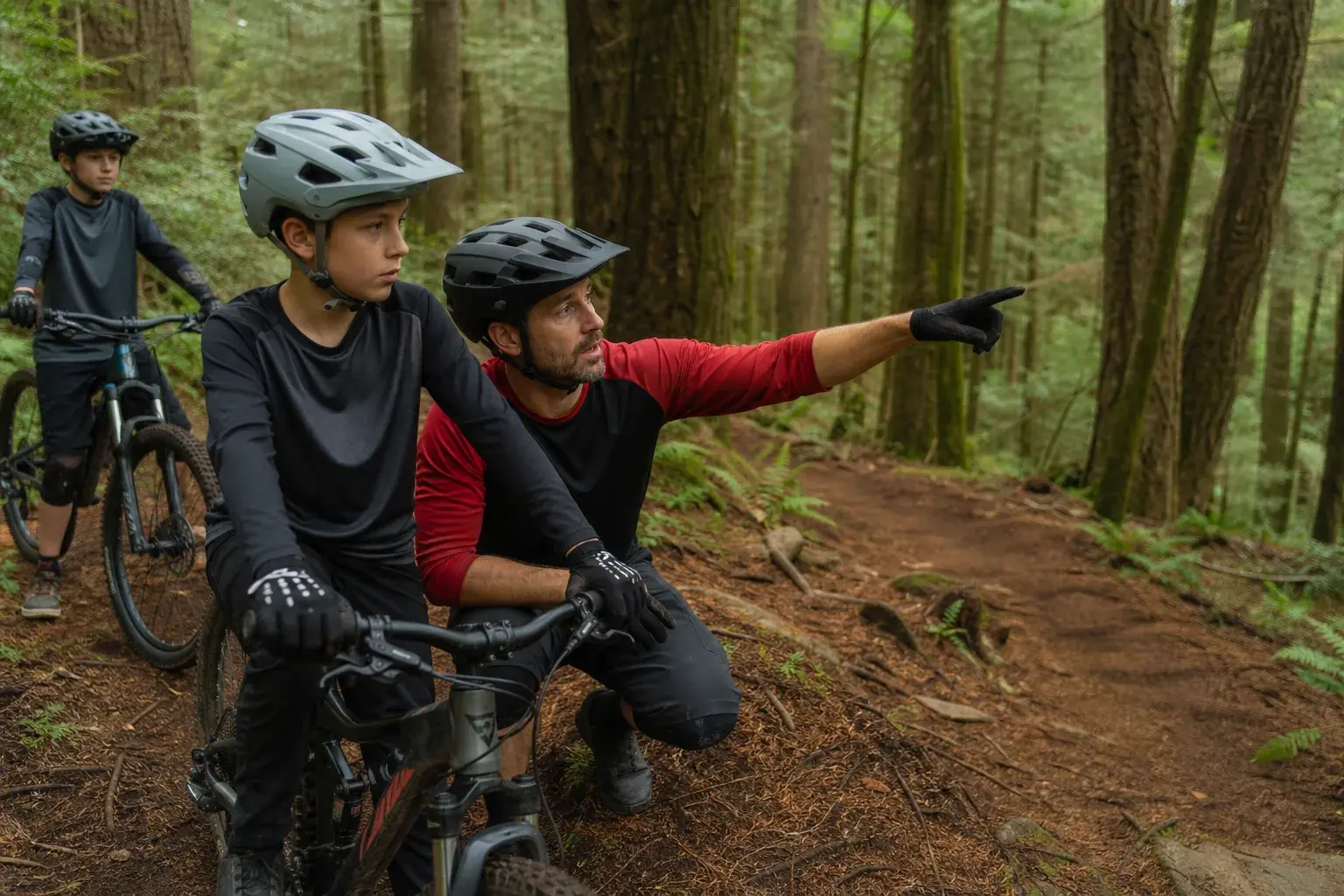 A mountain bike coach explaining line choice to a young rider beside a forest trail