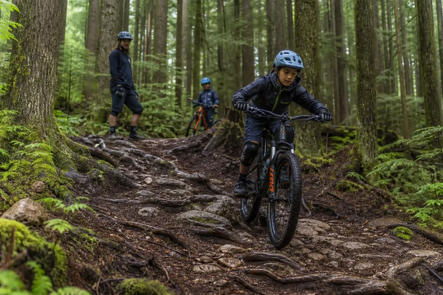 A young mountain biker chooses a controlled line through roots and rocks while a coach watches from beside the trail